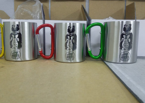 Four metal mugs with colorful carabiner handles on a wooden surface with laser engraving